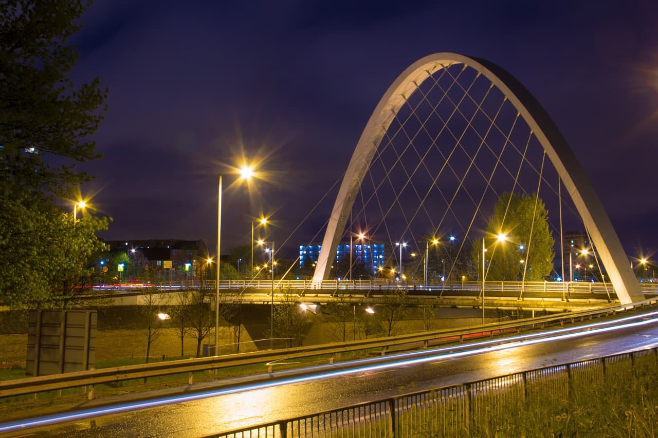A dual carriageway at night under a lit arch bridge, car lights drawn into long trails